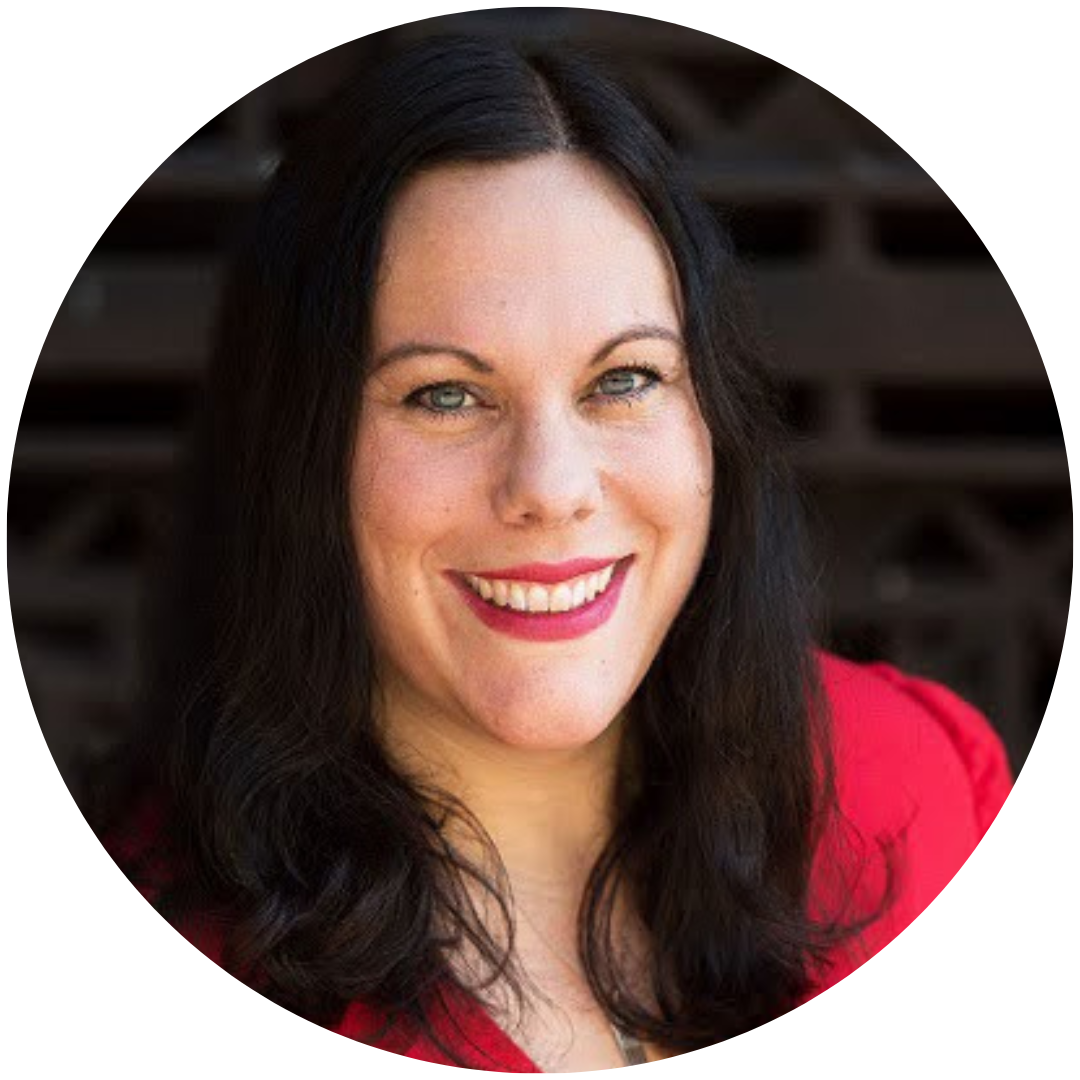 headshot image of a white woman with black hair and green eyes smiling at the camera in front of a brick wall. She is wearing a red top and red lipstick.