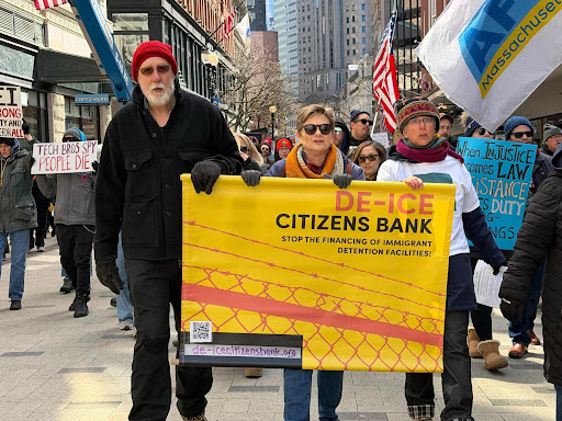 Three people participating in a march. They are carrying a large yellow banner that reads, 'De-ICE Citizens Bank. Stop the financing of immigrant detention facilities!'