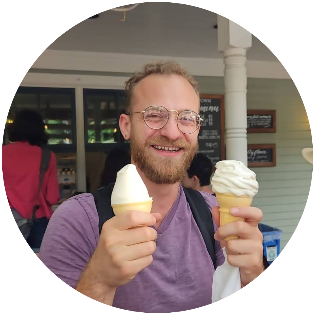 a person with short hair, glasses, and facial hair wearing a mauve shirt and holding two ice cream cones and smiling at the camera